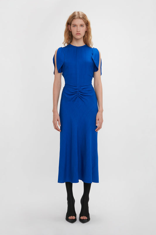 A woman in a Victoria Beckham palace blue midi dress with cap sleeves and a gathered waist, standing against a plain white background.