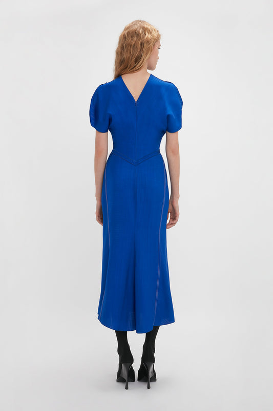 Woman in elegant Victoria Beckham Gathered Waist Midi Dress In Palace Blue and black heels standing with her back to the camera against a white background.