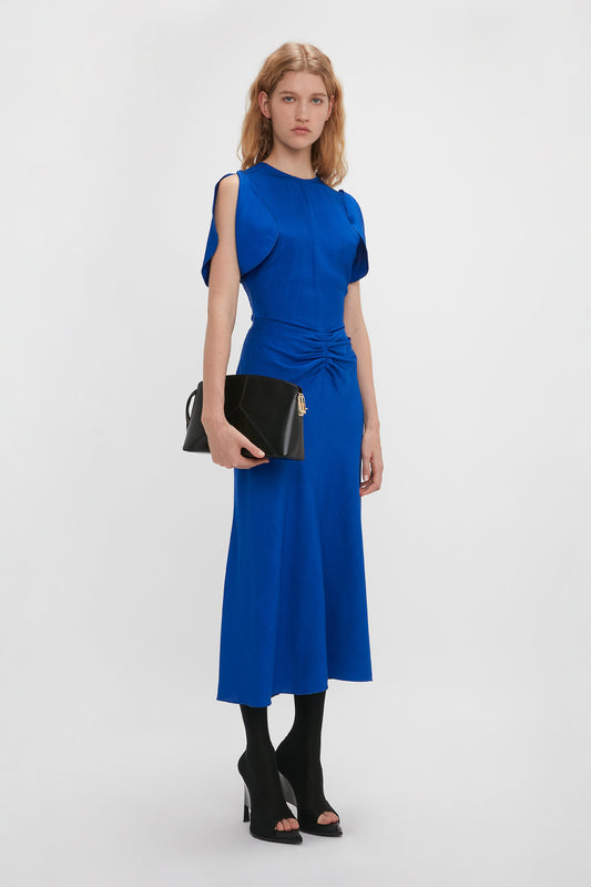 A woman in a bright Victoria Beckham Gathered Waist Midi Dress In Palace Blue with cap sleeves, holding a black clutch, stands against a white background. She wears black open-toe heels.