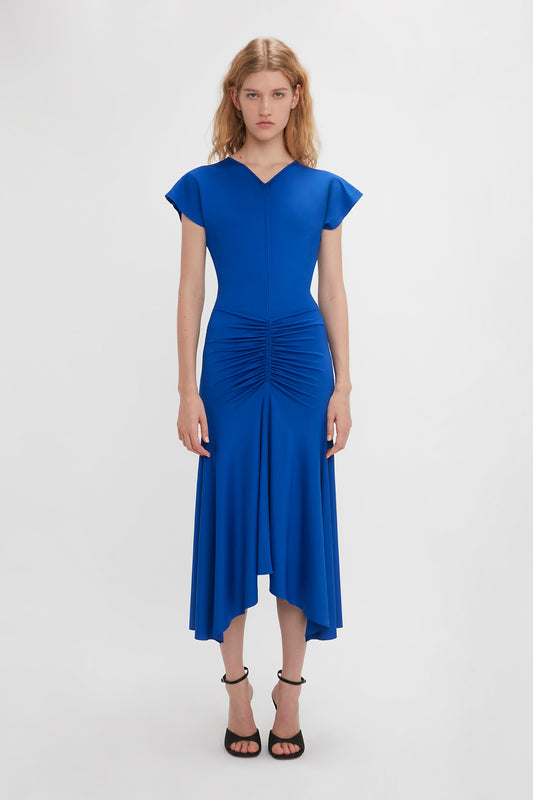 A woman in a bright royal blue Victoria Beckham mid-length dress with sleeveless and ruched details, standing against a white background. She wears black heeled sandals.