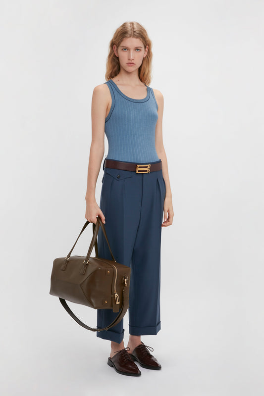 A woman in a blue fine knit tank top, Victoria Beckham wide leg cropped trousers in heritage blue, brown belt and shoes, holding a brown handbag against a white background.