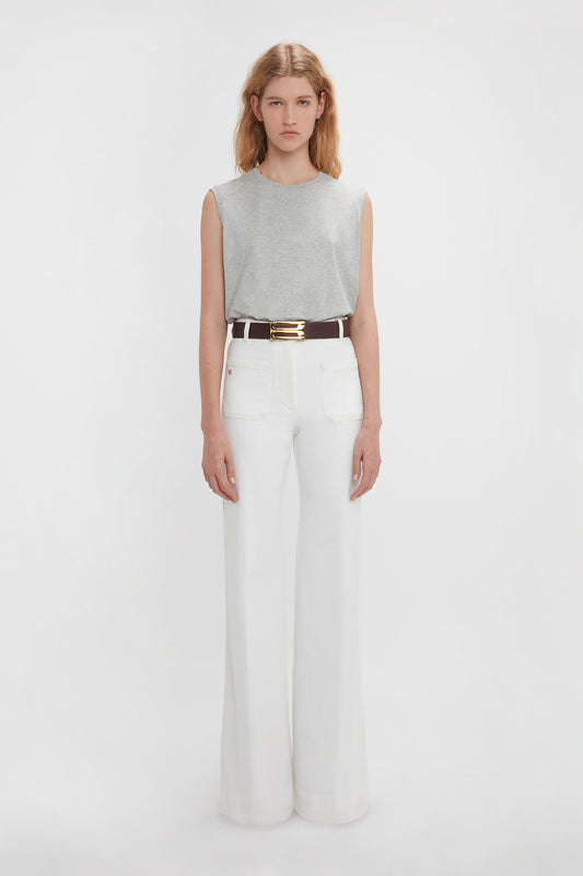 A person with long hair is standing against a plain background, wearing a Victoria Beckham Sleeveless T-Shirt In Grey Marl and high-waisted white trousers with a black belt.