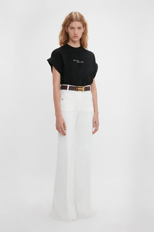 A young woman standing against a white background, wearing a Victoria Beckham 'Do As I Say, Not As I Do' Slogan T-Shirt in Black and white high-waisted flared jeans, accessorized with a slim belt.