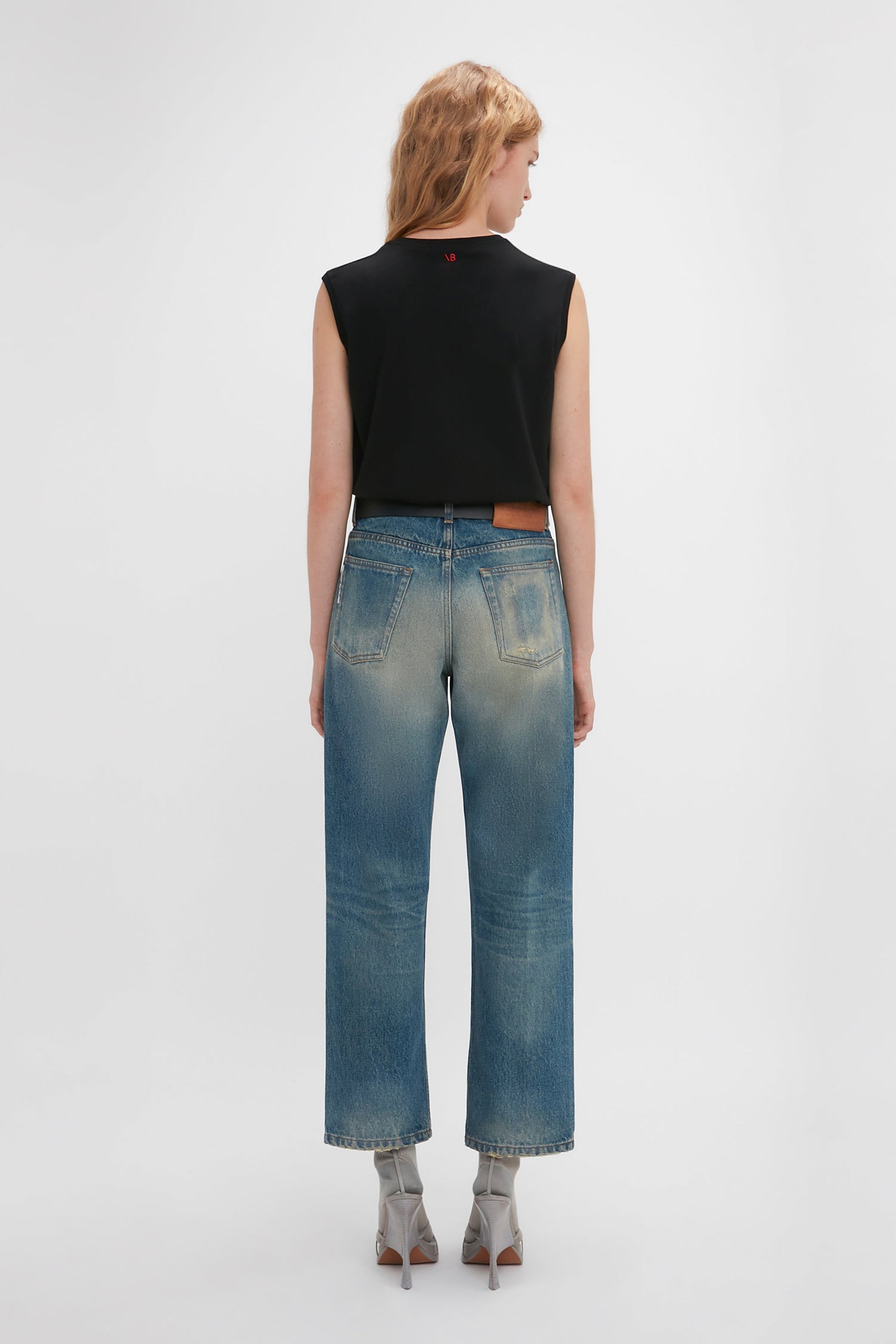 Rear view of a woman wearing a black sleeveless top and blue Victoria Beckham Relaxed Straight Leg Jean In Antique Indigo Wash, standing against a white background.