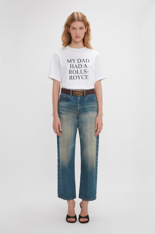 A woman stands against a plain background, wearing a relaxed fit Victoria Beckham slogan T-shirt with "my dad holds a Rolls-Royce" text, paired with faded jeans and black sandals.