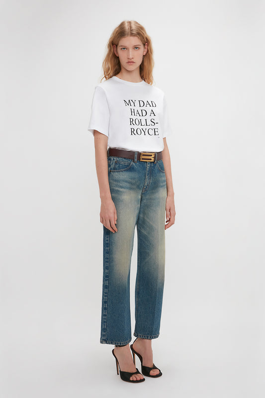 Young woman in a relaxed fit Victoria Beckham white t-shirt with the slogan "my dad has a rolls royce" and blue jeans, standing against a seamless white background.