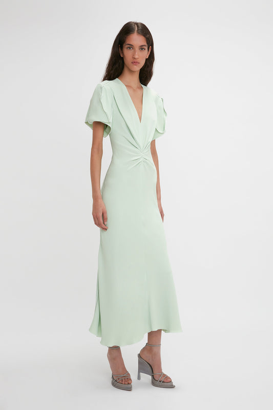 A woman in a light green Victoria Beckham Gathered V-Neck Midi Dress in Jade standing against a white background, the dress has short sleeves and waist-defining pleat detail.