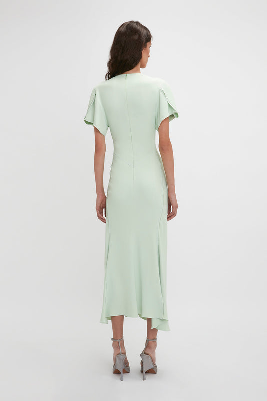 A woman from behind, wearing an elegant, pastel green Gathered V-Neck Midi Dress In Jade with flutter sleeves and a high collar, paired with light grey heels by Victoria Beckham.