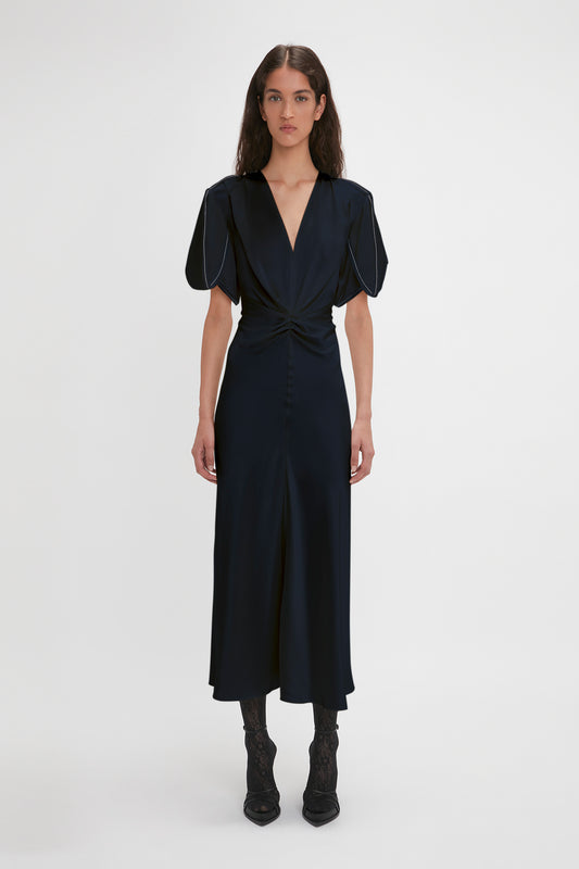 A woman in an Exclusive Gathered V-Neck Midi Dress In Navy by Victoria Beckham with puffed sleeves, contrasting topstitching, and cinched waist standing against a white background.