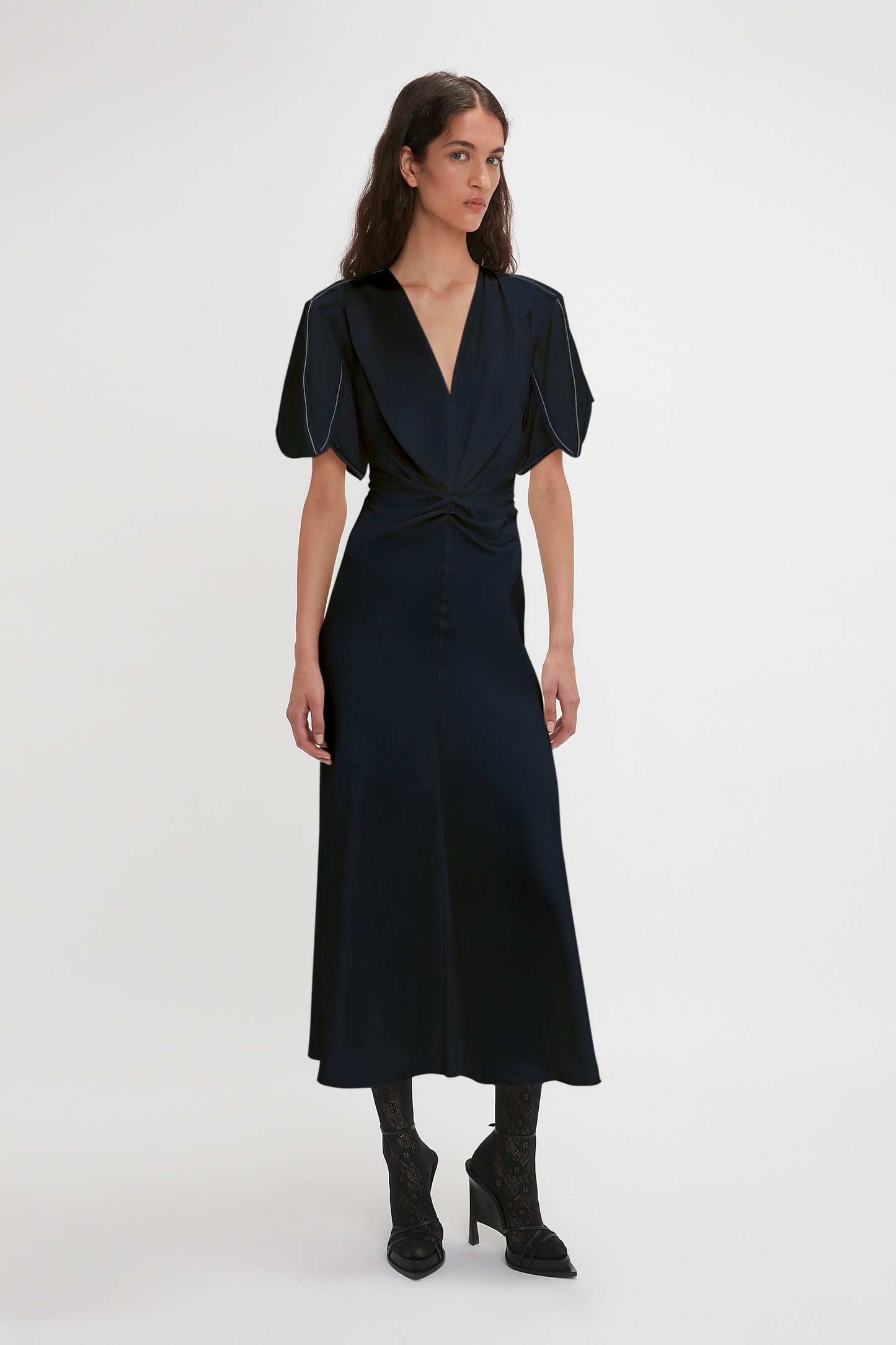 A woman wearing a Victoria Beckham navy blue Exclusive Gathered V-neck midi dress with puffed sleeves, stretch fabric, and black lace-up boots, standing in a studio with a white background.
