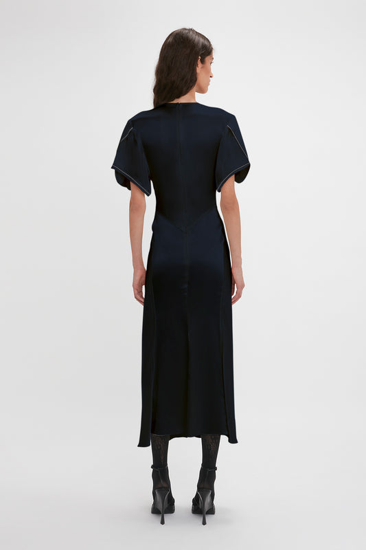 A woman in a Victoria Beckham Exclusive Gathered V-Neck Midi Dress In Navy with waist-defining pleat detail and black heeled boots stands facing away from the camera on a white background.
