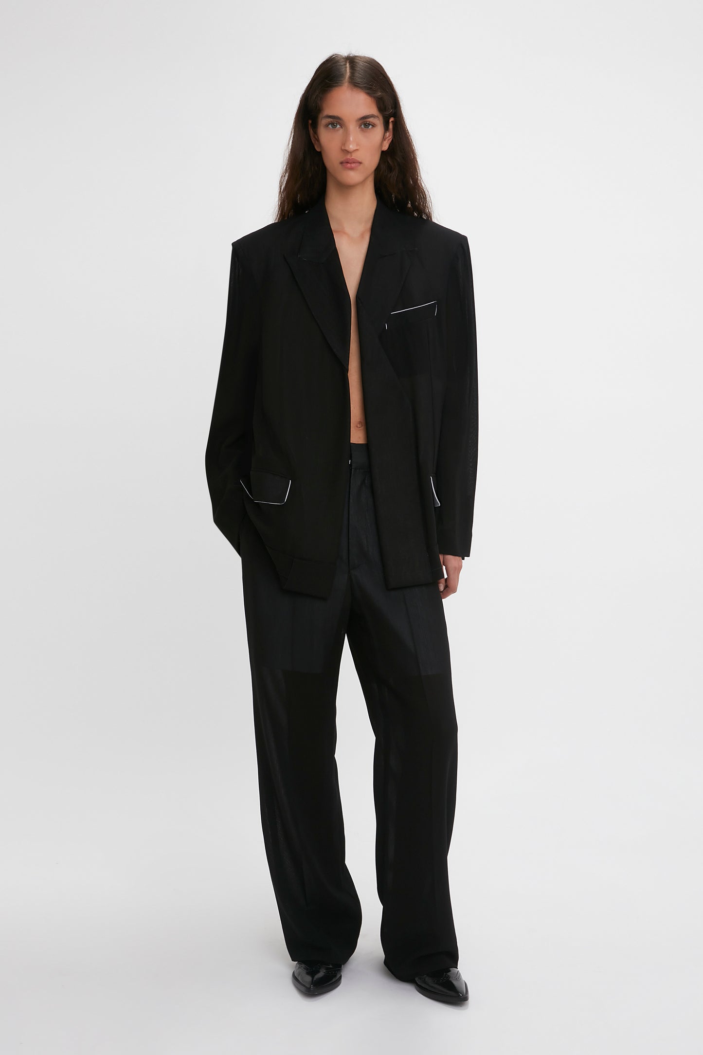 A woman in a sleek black suit with a Victoria Beckham Fold Detail Tailored Jacket featuring folded detail and straight leg trousers, standing against a plain white background.