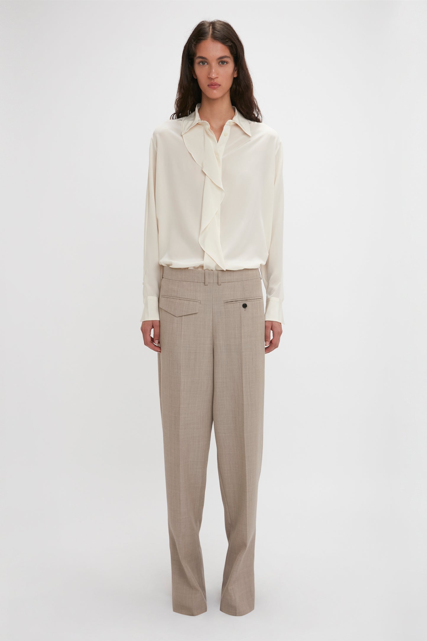 A person stands against a plain background, wearing the Victoria Beckham Asymmetric Ruffle Blouse In Ivory with voluminous sleeves and a ruffled front, paired with beige trousers featuring a button-front closure.