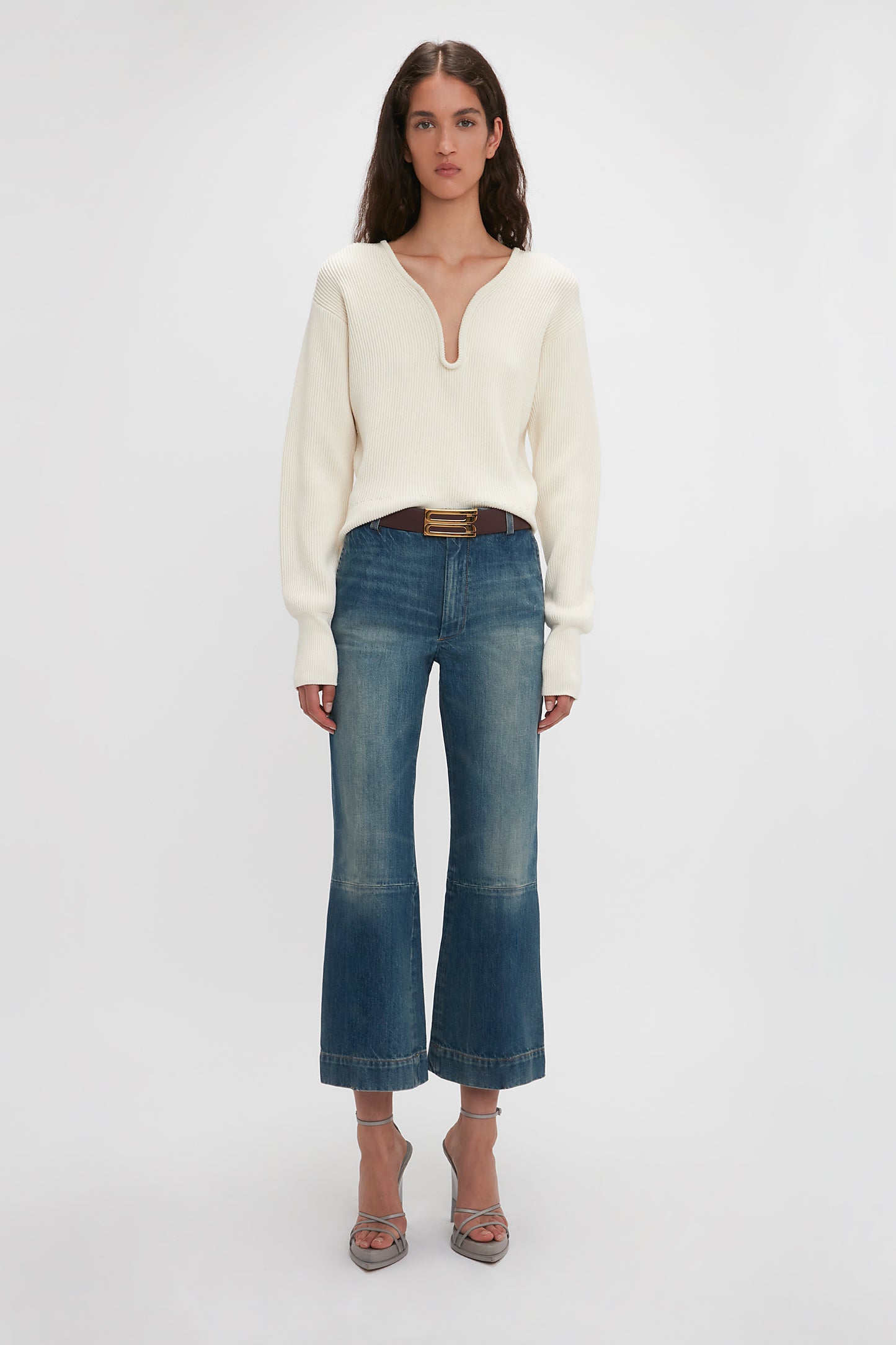 A woman wearing a white v-neck sweater and Victoria Beckham's Cropped Kick Jean In Indigrey Wash standing against a plain white background.