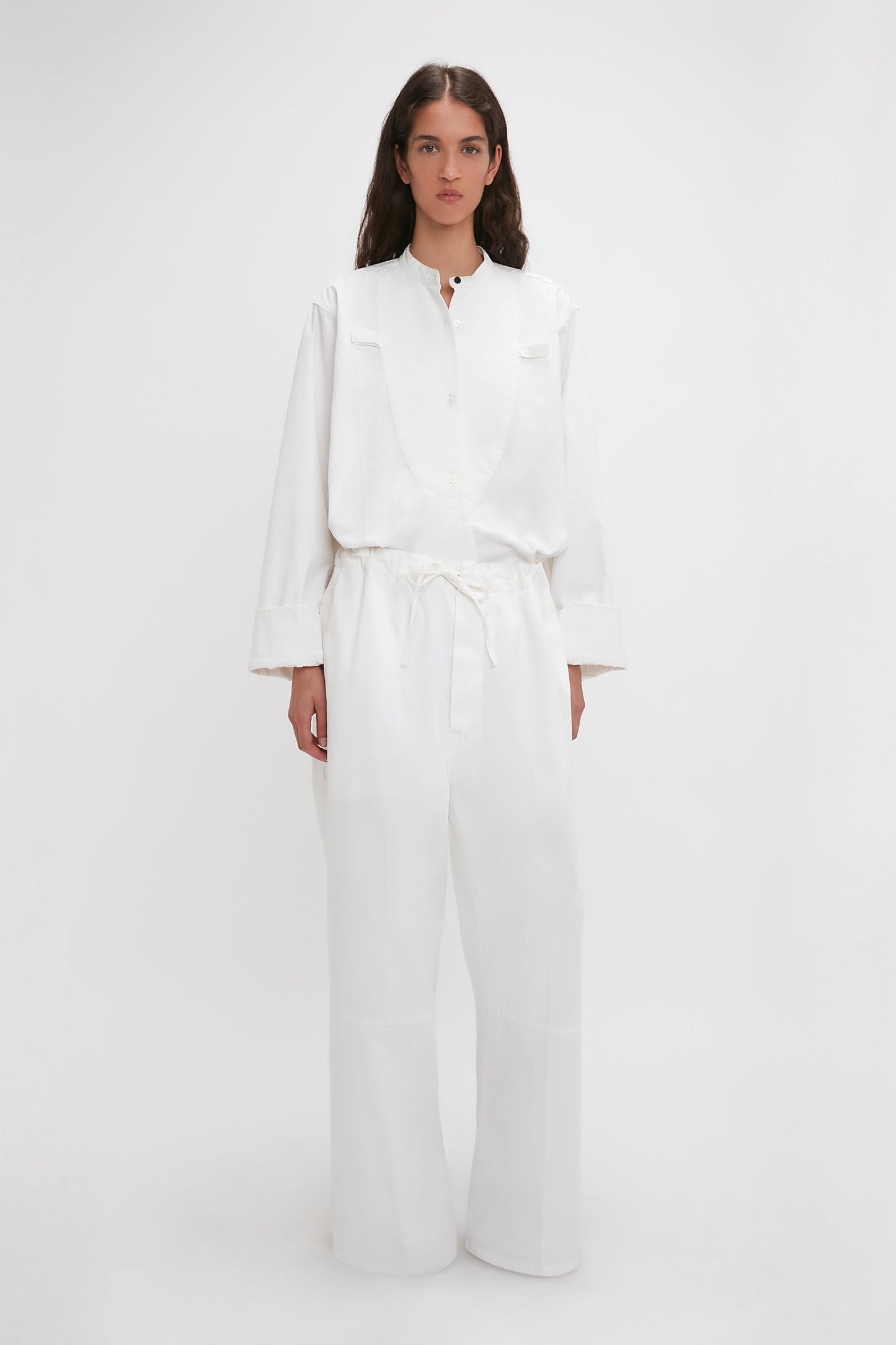 A woman in Victoria Beckham's drawstring pyjama trousers in washed white stands against a plain background, facing forward with a neutral expression.