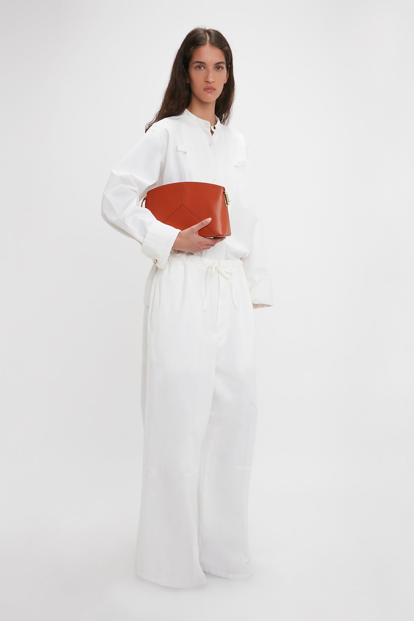 Person in a white outfit holding a Victoria Beckham Victoria Clutch Bag In Tan Leather against a plain white background.