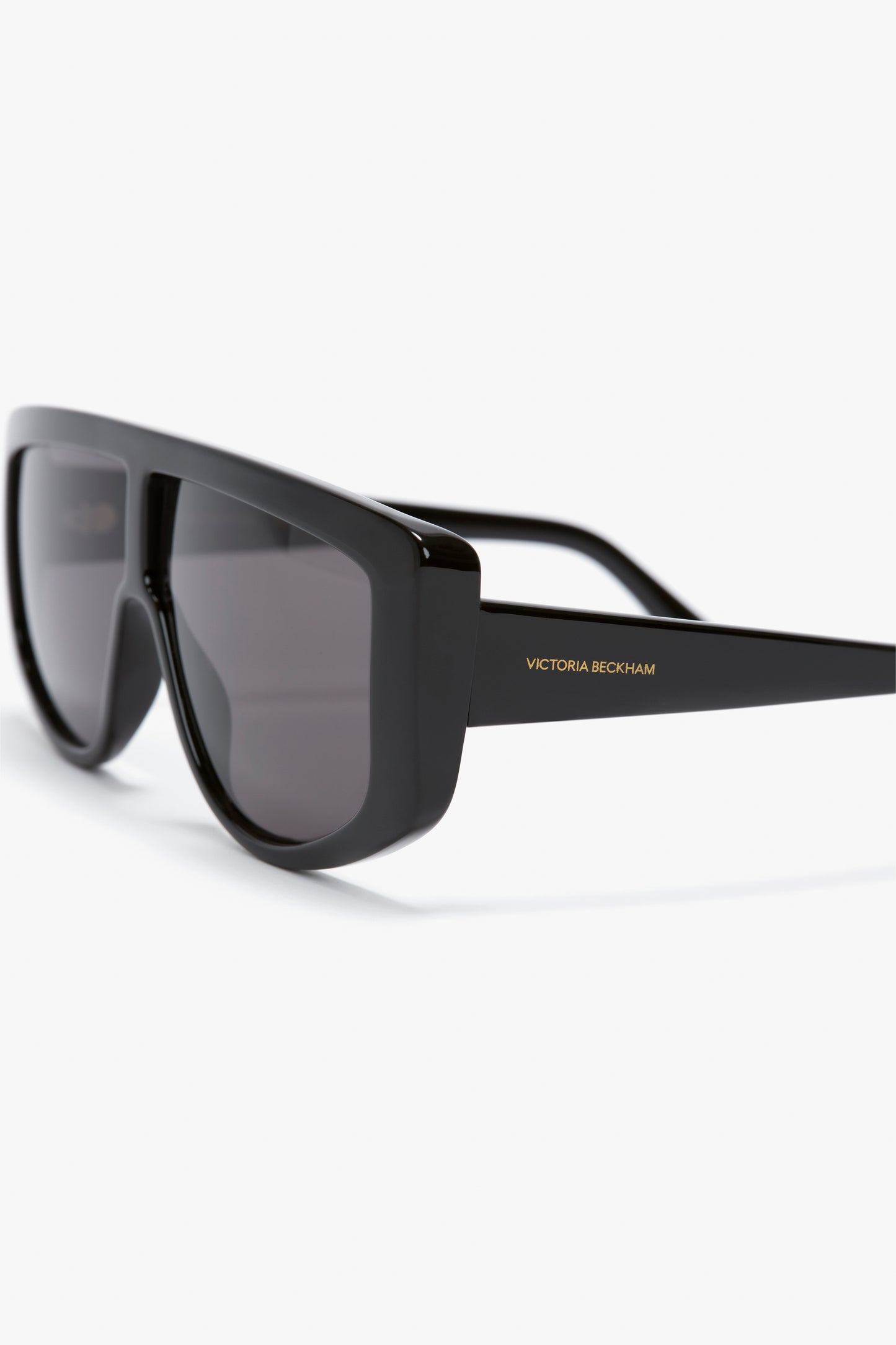 Close-up of Acetate Visor Sunglasses In Black with thick, oversized frames. The brand name "Victoria Beckham" is printed on the temple, and the black tonal lenses complete the chic look of these Victoria Beckham eyewear pieces.