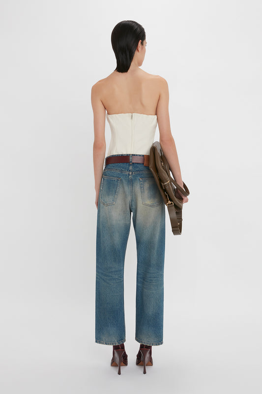 A woman seen from the back wearing a strapless Victoria Beckham Corset Top in Antique White, blue jeans, brown heels, and holding a brown handbag.