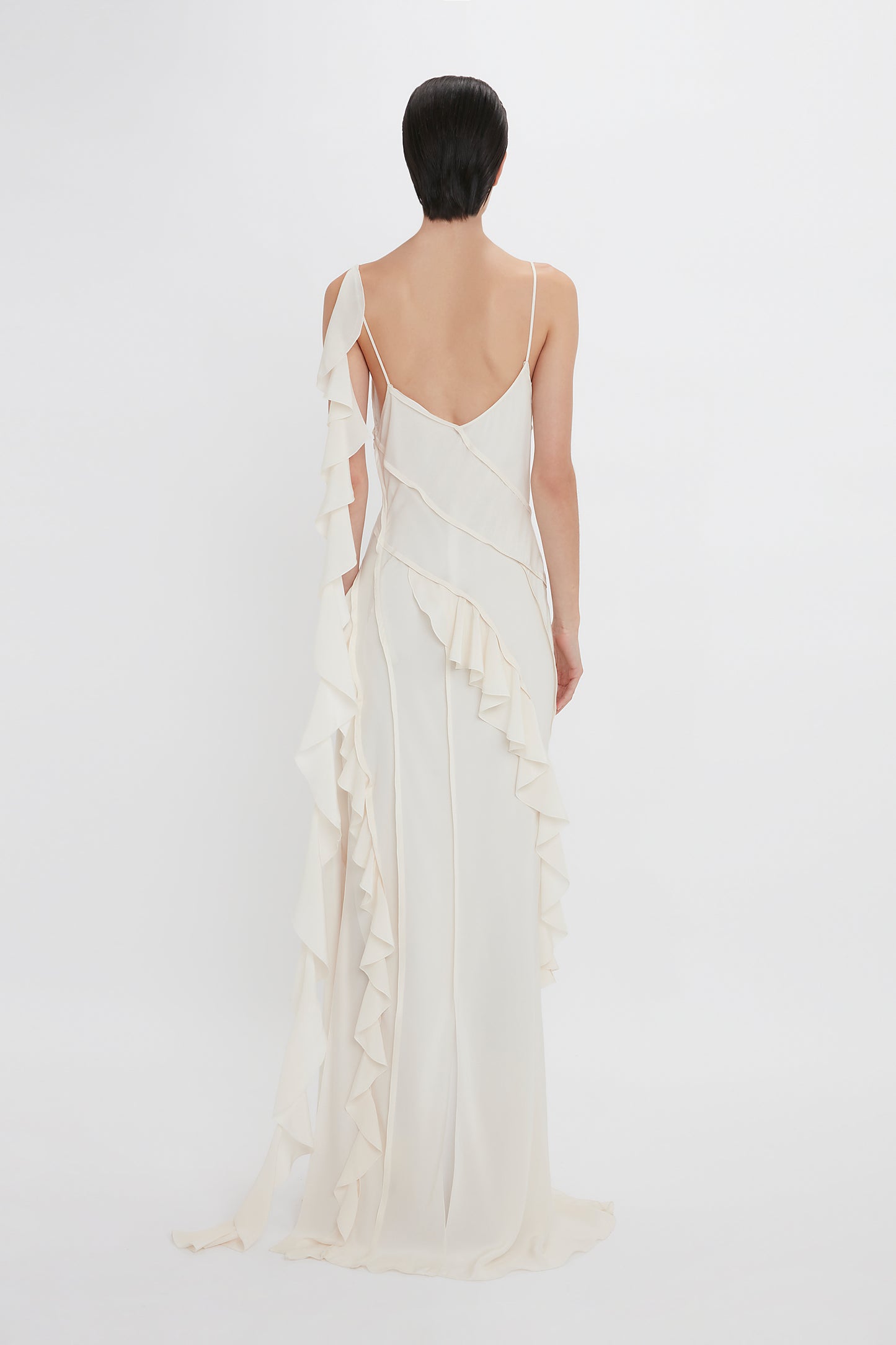 A woman in an Exclusive Asymmetric Bias Frill Dress In Ivory by Victoria Beckham, with lace detailing on the back, standing against a white background.