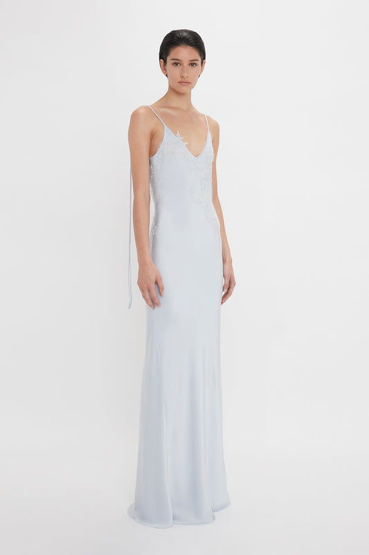 A woman in a Victoria Beckham Exclusive Lace Detail Floor-Length Cami Dress In Ice with delicate lace detailing on the bodice, standing against a white background.