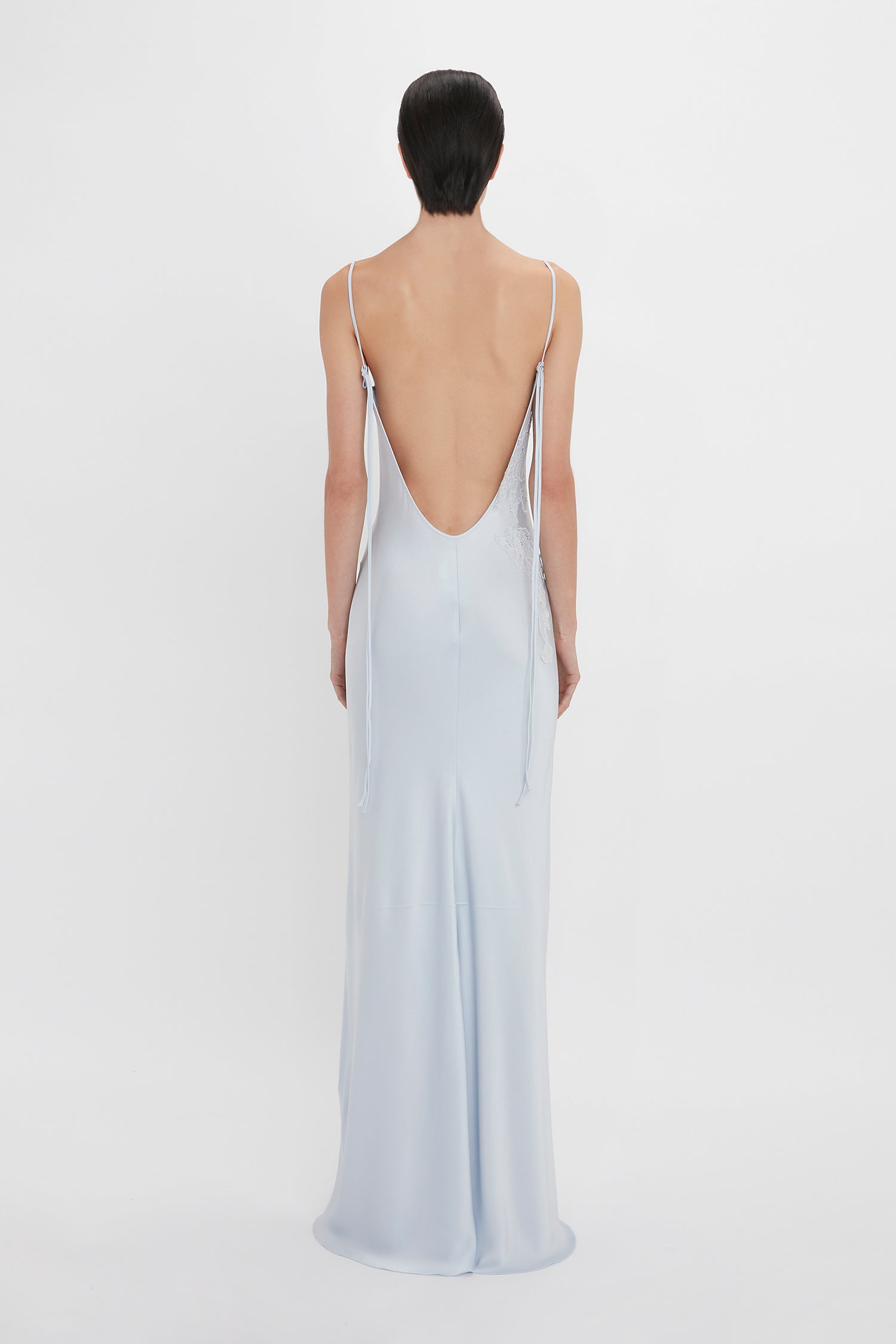 A woman facing away wearing a Victoria Beckham Exclusive Lace Detail Floor-Length Cami Dress In Ice, with a low-cut back and thin straps, on a plain white background.