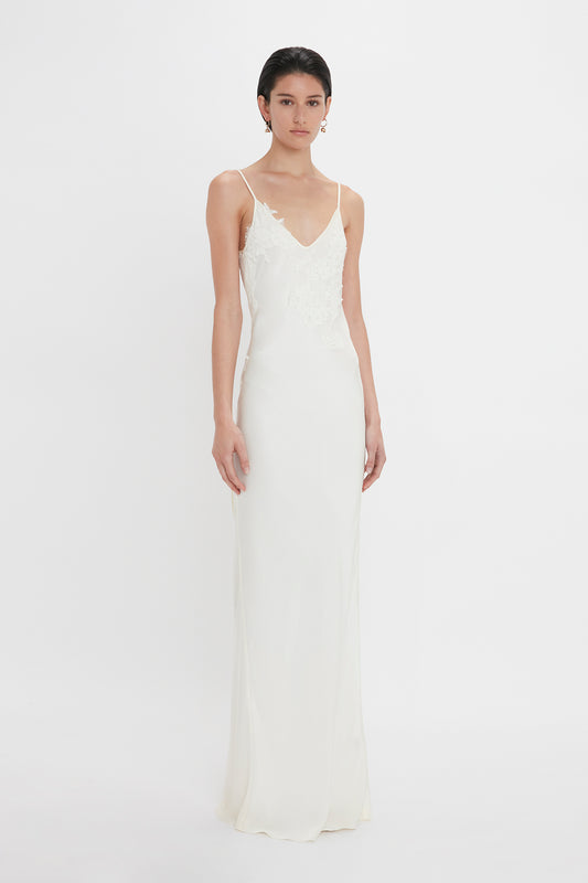 A woman in a sleek, sleeveless white wedding dress with lace detailing on the bodice, accessorized with Victoria Beckham's Exclusive Camellia Flower Hoop Earrings In Gold, standing against a plain white background.