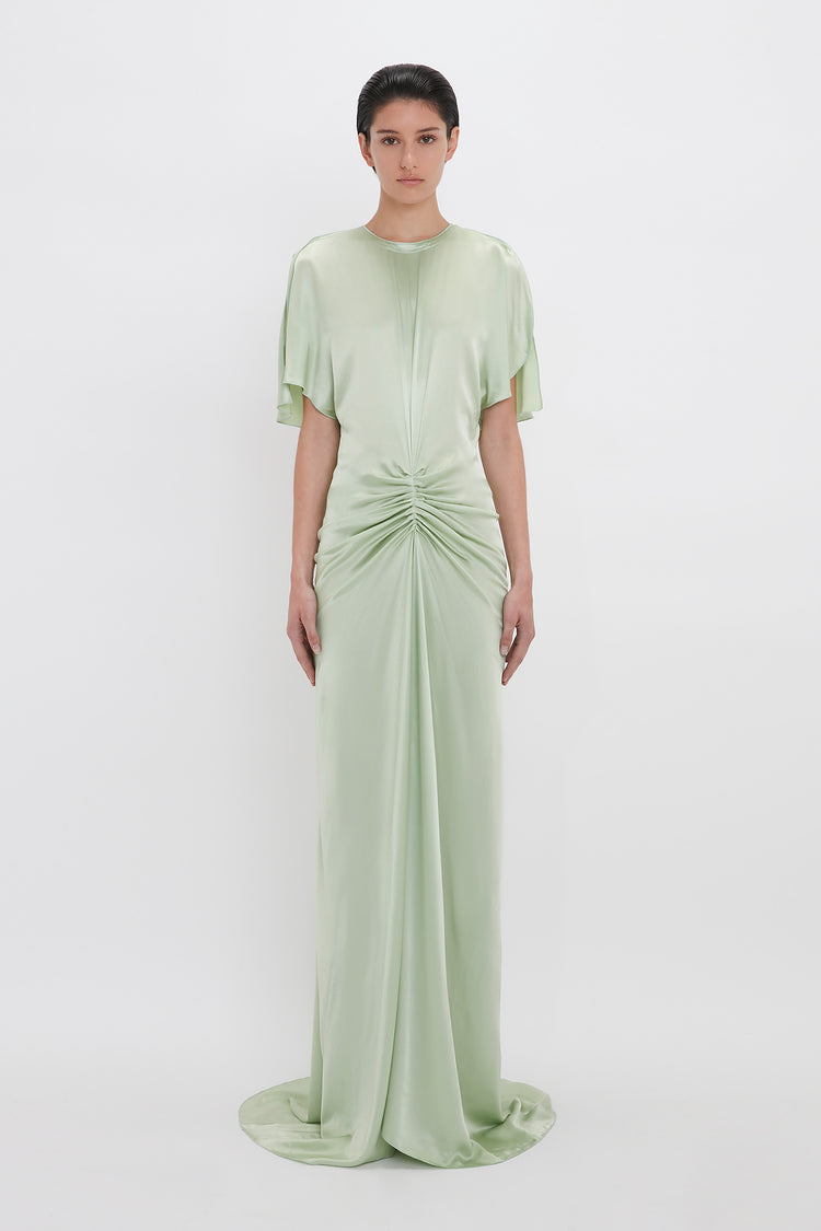 A woman models a pale green, elegant, Exclusive Floor-Length Gathered Dress In Jade by Victoria Beckham with a gathered twist at the waist and short sleeves, standing against a white backdrop.