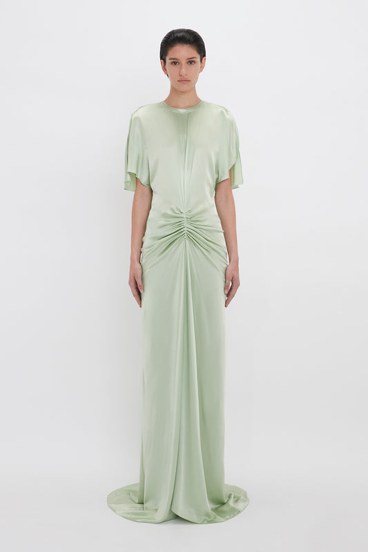 A woman models a pale green, elegant, Exclusive Floor-Length Gathered Dress In Jade by Victoria Beckham with a gathered twist at the waist and short sleeves, standing against a white backdrop.