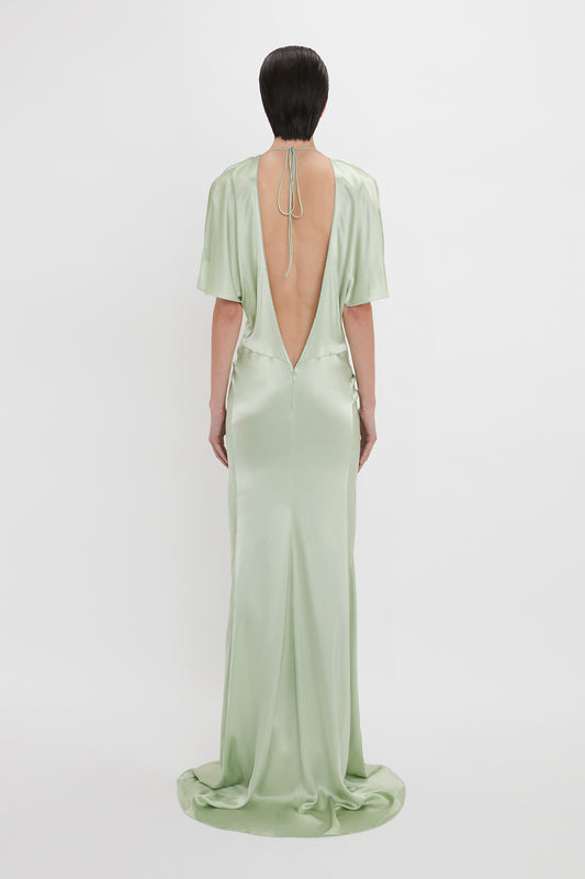 A woman stands with her back to the camera, wearing an elegant, light green, open back Victoria Beckham gown with a deep v-cut and a floor-length hem.