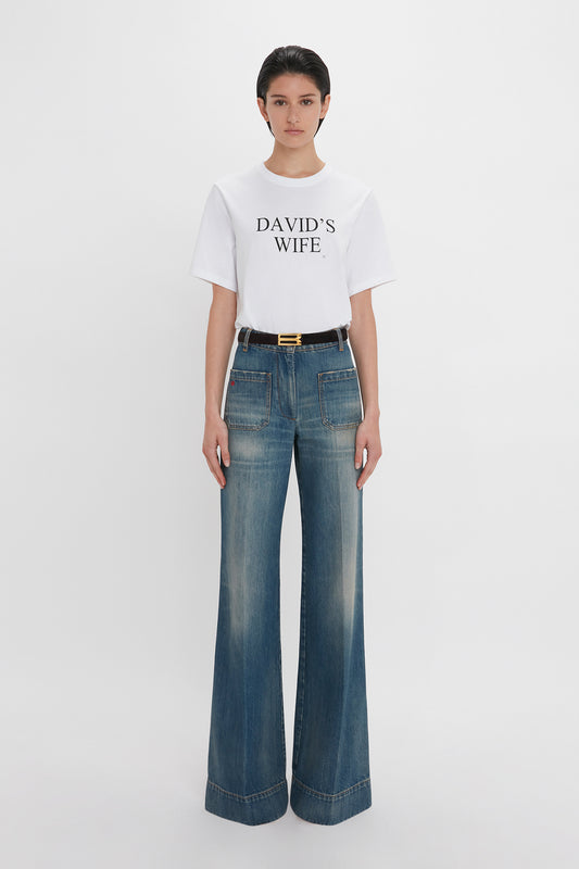 A woman wearing a "David's Wife" slogan T-shirt in white by Victoria Beckham and blue wide-legged jeans with a black belt stands against a white background.