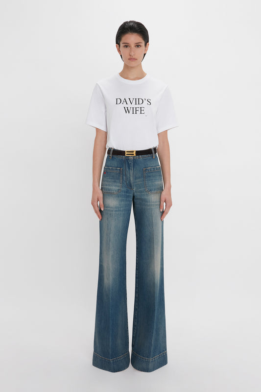 A person standing against a white background, wearing a white T-shirt with the text "DAVID'S WIFE" and blue Alina High Waisted Jean In Indigrey Wash by Victoria Beckham.