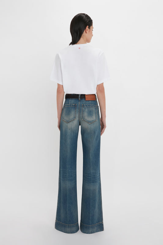 Rear view of a woman wearing a Victoria Beckham 'David's Wife' Slogan T-Shirt In White and flared blue jeans, standing against a plain white background.
