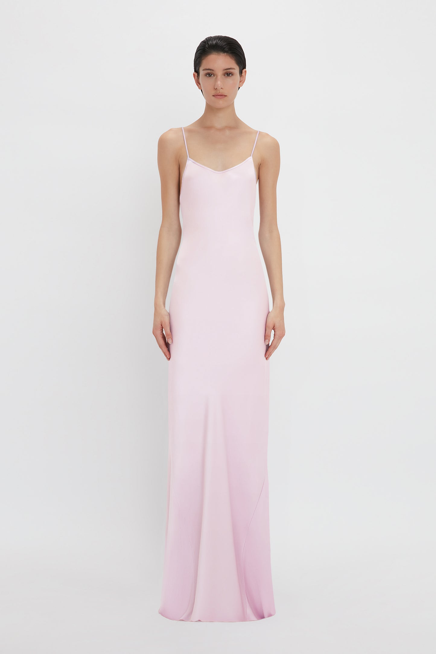 A woman stands against a white background wearing a long, light pink Low Back Cami Floor-Length Dress In Rosa by Victoria Beckham with thin straps.