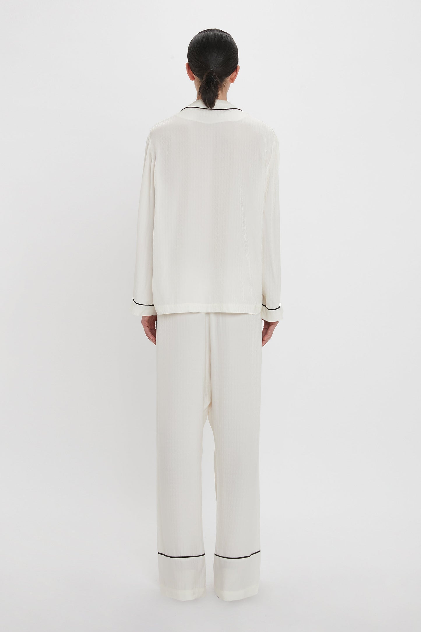 Person standing facing away, wearing a Victoria Beckham VB Monogram Pyjama Sleep Set In Ivory, an all-white loungewear outfit with black trim, including a long-sleeved top and loose-fitting pants. 100% silk fabric creates a luxurious feel. The background is plain white.
