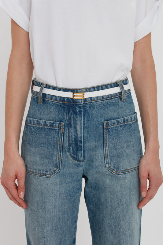 Close-up of a person wearing blue jeans and a white t-shirt, with a focus on a Victoria Beckham Exclusive Micro Frame Belt In White Leather with gold hardware.