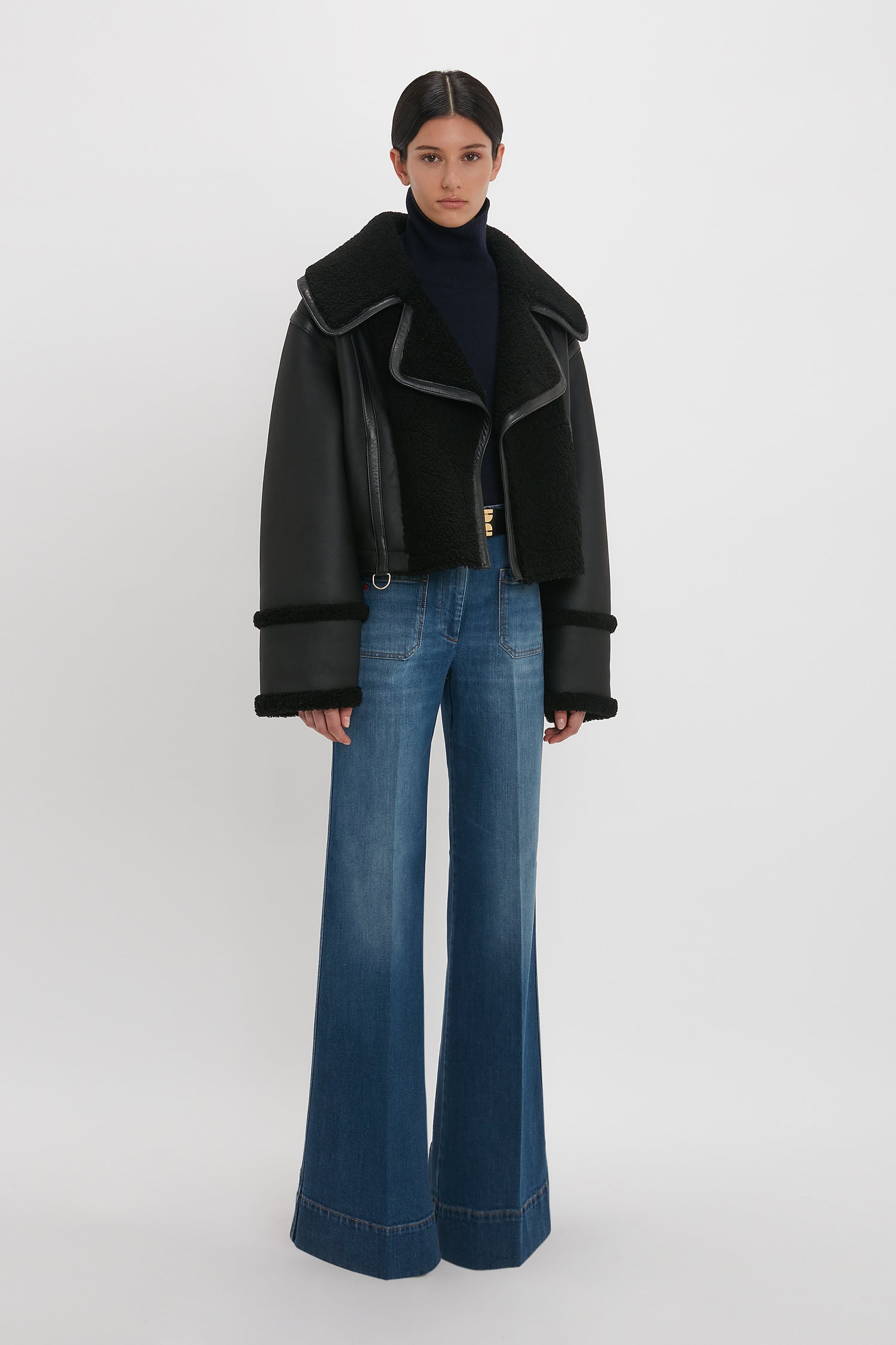 A person stands wearing a luxurious Shearling Jacket In Black by Victoria Beckham, navy turtleneck, and wide-leg blue jeans. The background is plain white.