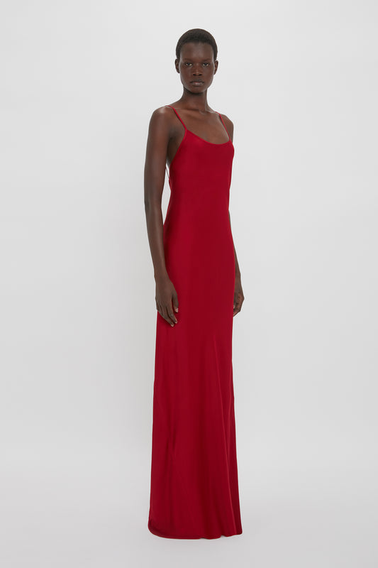 A person with short hair is wearing a long, flattering fit Low Back Cami Floor-Length Dress In Poppy Red by Victoria Beckham with thin straps, posing against a plain white background.