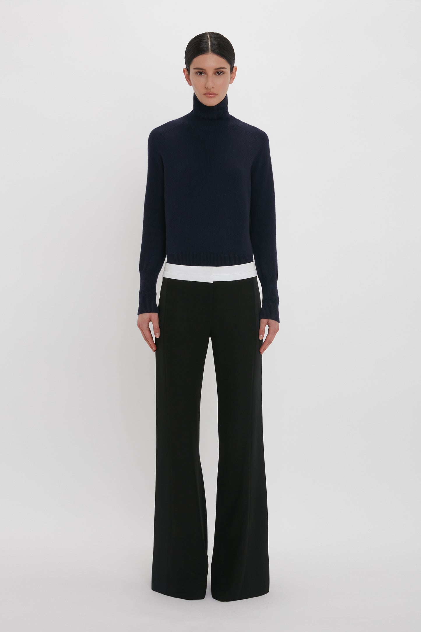 A person stands against a plain white background wearing a dark turtleneck sweater and Victoria Beckham's Side Panel Trouser In Black.