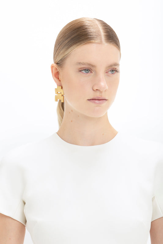A woman with blonde hair slicked back, wearing a Victoria Beckham fitted T-shirt dress in ivory and a large gold earring, against a plain white background.