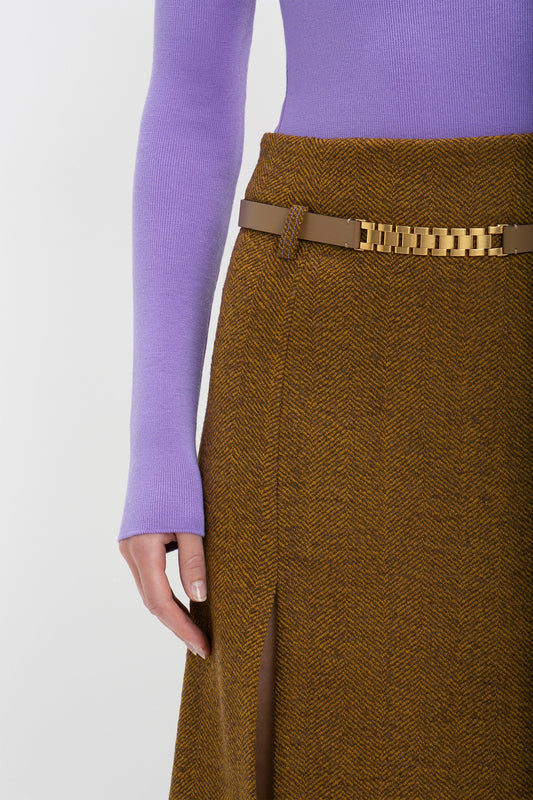 Close-up view of a person wearing a purple sweater and a textured brown skirt with a golden chain belt, accessorized with a Victoria Beckham calf-leather Watch Strap Detail Belt in Khaki-Brown.