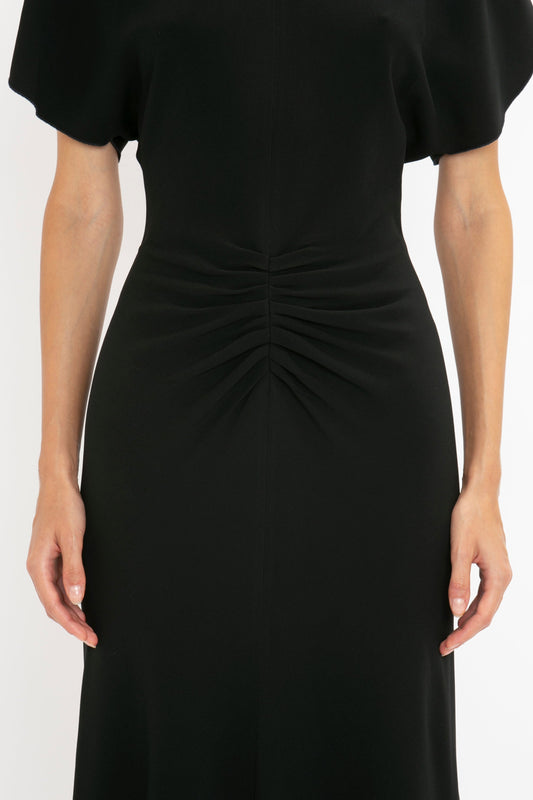 Close-up of a woman in a black dress with ruched detailing at the waist and flutter sleeves, focusing on the midsection of her Victoria Beckham Gathered Waist Midi Dress in Black.