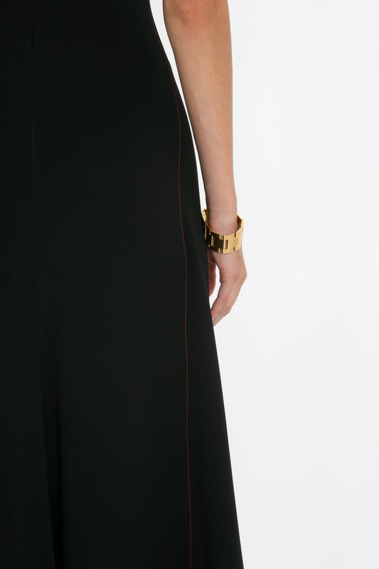 A close-up view of a woman's side, focusing on her wrist wearing a gold bracelet, against a Victoria Beckham black dress with draped front-tie and subtle red stitching.