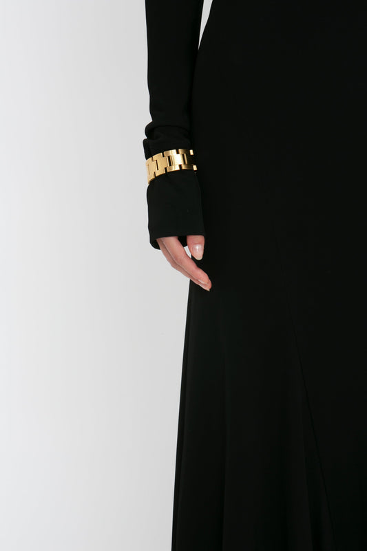 Close-up of a woman's arm in a Victoria Beckham black gown with a gold bracelet, against a white background.