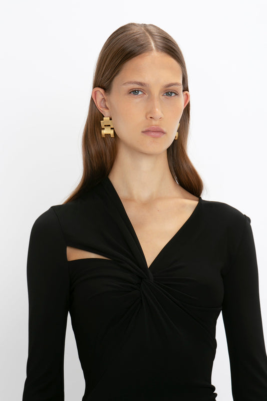 Woman wearing a Victoria Beckham Tie Detail Floor-Length Dress in Black and geometric earrings, standing against a white background.