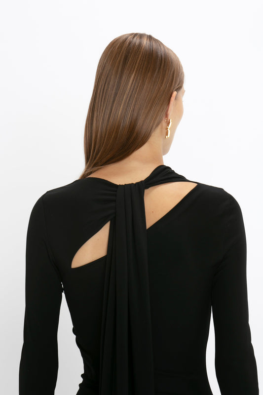 Woman in a Victoria Beckham Tie Detail Floor-Length Dress in Black with a unique asymmetric cut-out back design, viewed from behind against a white background.