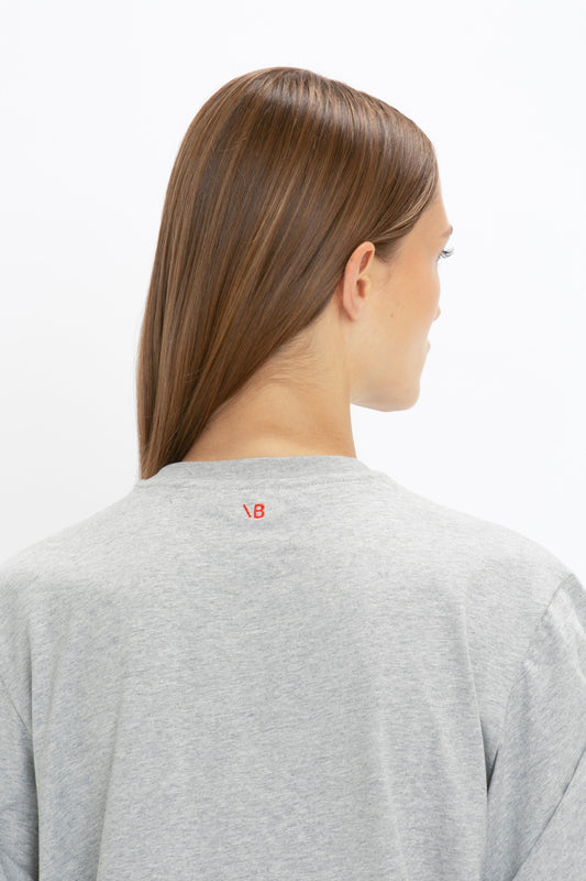 Profile view of a woman wearing an Asymmetric Relaxed Fit T-Shirt In Grey Marl made of organic cotton by Victoria Beckham, with a small red logo on the back, facing away from the camera.