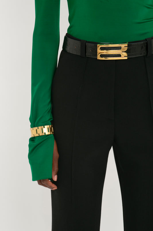 Close-up of a person wearing black pants and a green top with a Victoria Beckham Jumbo Frame Belt in Black Croc-Effect Leather and bracelet, focusing on the midsection and arm.