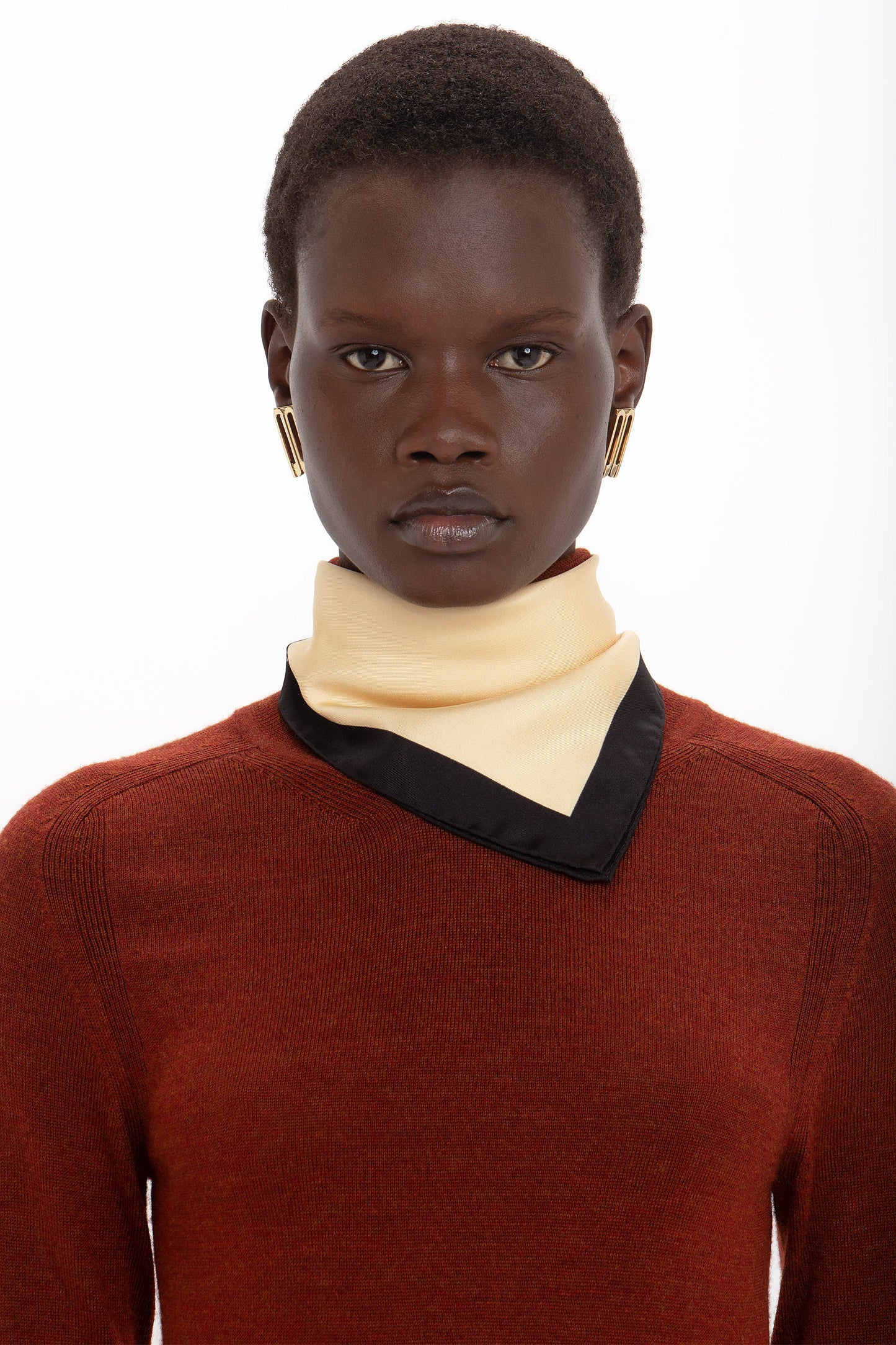 Person wearing a red sweater, a luxurious Colour Block Silk Scarf In Macadamia by Victoria Beckham, and gold rectangular earrings, looking directly at the camera against a white background.