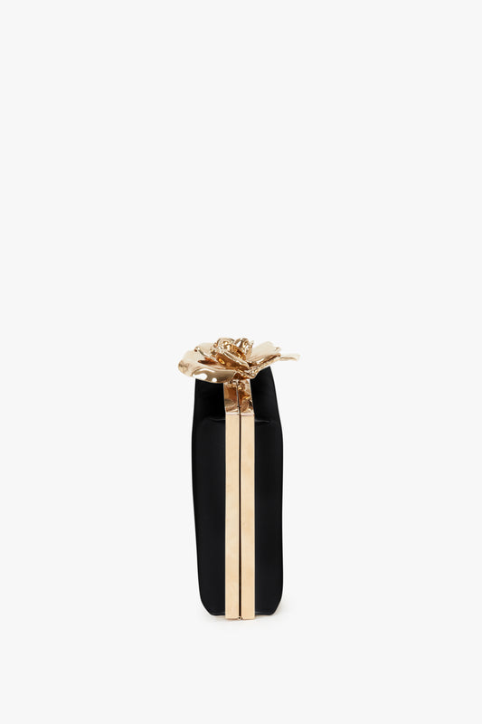 A black and gold cylindrical Victoria Beckham Frame Flower Minaudiere adorned with a shiny gold flower clasp, isolated on a white background.