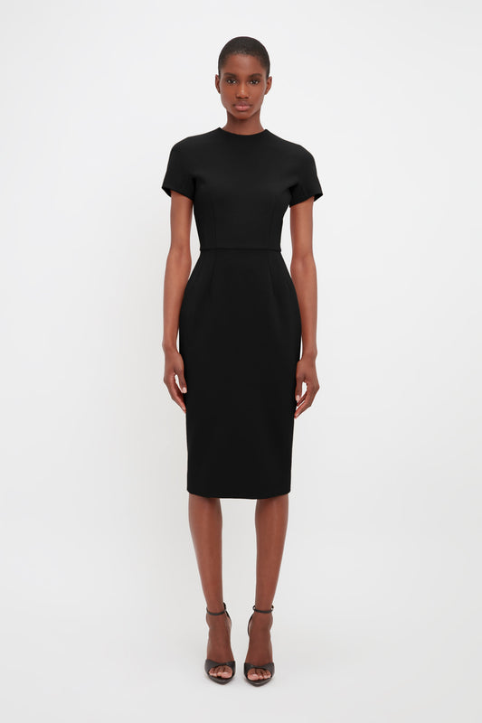 A woman in a Victoria Beckham fitted black knee-length dress made of bonded crepe fabric stands against a plain white background, looking directly at the camera.